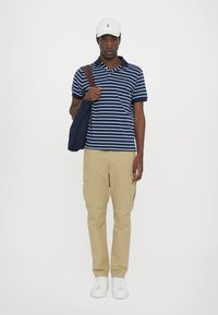 Cămașă polo cu dungi albastre și albe, pantaloni cargo khaki, adidași albi și o șapcă albă, cu o geantă de umăr marron pe umărul stâng.
