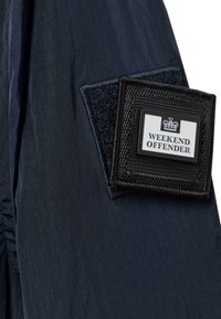 Die marineblaue Stoffjacke hat ein strukturiertes schwarzes Patch mit dem weißen Aufdruck "WEEKEND OFFENDER", das mit Klettverschluss am Ärmel befestigt ist.