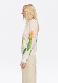 Lichtbeige sweatshirt met bloemontwerpen in oranje en groen. Heeft een relaxed fit en ribgebreide cuffs. Geddragen met lichtbeige broek.
