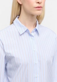 Donna con capelli biondi lisci, che indossa una camicia button-up azzurro chiaro con sottili righe verticali rosa e bianche.