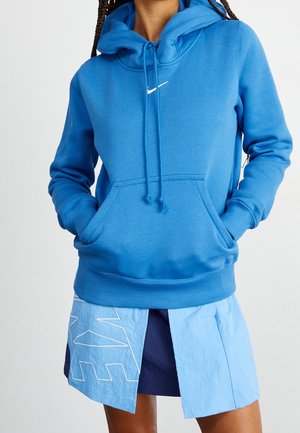 Sweat à capuche Nike en bleu vif, avec une poche frontale, une capuche à cordon et un logo blanc, associé à une jupe bleue superposée.