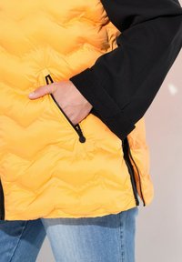 Daunenjacke mit leuchtend gelbem, gestepptem Außenmaterial und schwarzen Ärmeln; verfügt über eine seitliche Reißverschlusstasche und eine glatte Oberfläche.