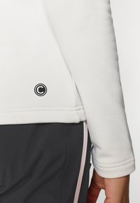 Top blanco de manga larga con un ajuste ceñido, presenta un logo negro circular cerca del dobladillo, complementado por pantalones deportivos grises con acentos rosas.