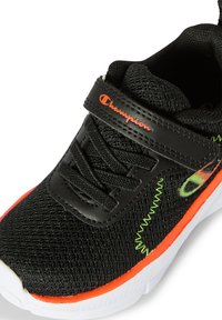 Scarpa sportiva nera con tomaia in rete, fascia regolabile, suola bianca e accenti arancioni e verdi lungo il lato. Dettaglio della superficie testurizzata.