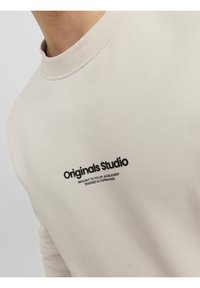 Gros plan sur le col d'une chemise beige avec le texte noir « Originals Studio » et un texte plus petit en dessous sur la poitrine.