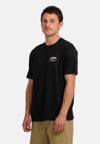 Zwarte katoenen T-shirt met korte mouwen, ronde hals, en een logo patch op de borst. Model staat voor een eenvoudige witte achtergrond.