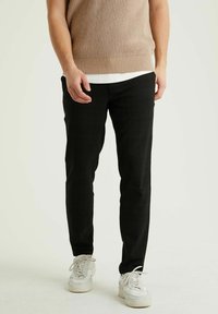 Muž v černých slim-fit kalhotách, béžovém pleteném svetru přes bílou košili a bílých teniskách, stojící na pozadí jednoduchého světlého pozadí.