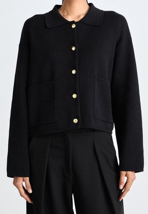 Czarny kardigan z długim rękawem z dzianiny z złotymi guzikami i przednimi kieszeniami, zestawiony z luźnymi czarnymi spodniami na osobie.
