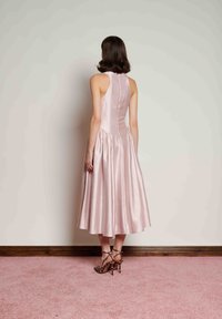 Φόρεμα από ελαφρύ ροζ σατέν με εφαρμοστό μπούστο και φαρδιά φούστα, που διαθέτει ψηλή πλάτη και ρυτιδωμένες λεπτομέρειες. Συνδυάζεται με ψηλοτάκουνα παπούτσια λεοπάρ.