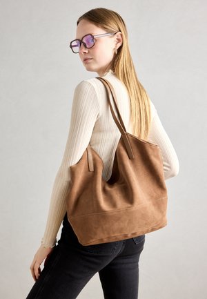 Femme aux longs cheveux blonds portant des lunettes et un haut blanc côtelé porte un grand sac à bandoulière en daim marron sur un fond uni.