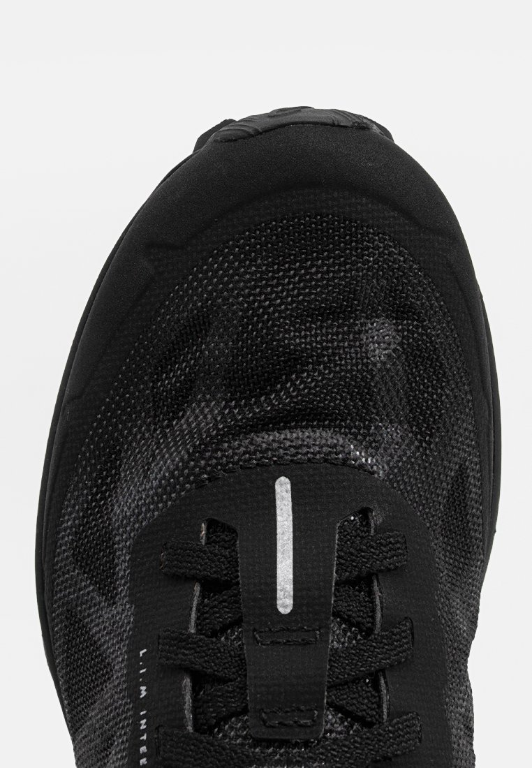 Zapatilla deportiva negra con parte superior de malla texturizada, cierre con cordones y acentos reflectantes. Presenta una puntera redondeada y suela acolchada.