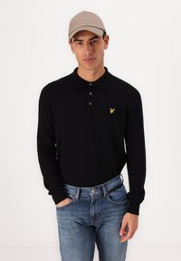 Polo negru cu mânecă lungă, cu trei butoane, având un mic logo cu o pasăre galbenă; purtat cu blugi albaștri și o șapcă bej deschis.