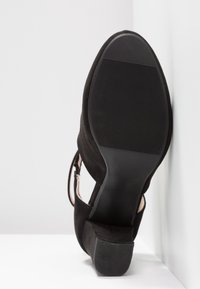 Chaussure à talon haut en daim noir avec une finition lisse, un design ouvert, et une semelle en caoutchouc noir texturée. Présente des lanières fines couleur camel.
