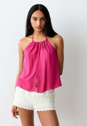 Femme aux longs cheveux foncés porte un haut rose sans manches à col halter avec une fente à l'avant et un short en dentelle blanche, debout devant un fond uni.