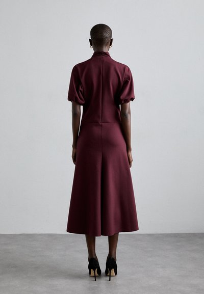 Rochie midi bordeaux cu mâneci scurte tip puff, guler înalt, bust ajustat și fustă evazată. Pantofi negri cu toc înalt completează look-ul.