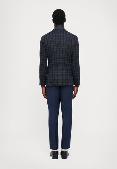Sacou bleumarin cu carouri, cu siluetă bine definită, talie strânsă și croială specifică, asortat cu pantaloni bleumarin eleganți și pantofi negri.