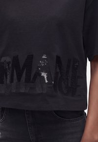 Μαύρο cropped t-shirt με λαμπερά μαύρα παγιετένια γράμματα που σχηματίζουν τη λέξη "ARMANI EXCHANGE" στο κάτω μέρος μπροστά, φορεμένο με μαύρο τζιν.