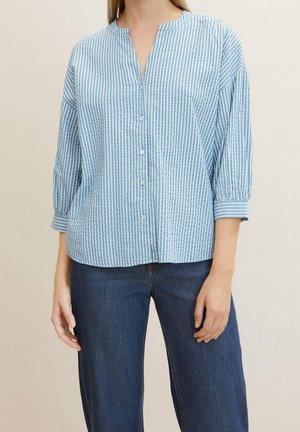 Kobieta nosząca jasnoniebieską i białą pionowo paskowaną koszulę zapinaną na guziki z rękawami o długości do łokcia oraz ciemnoniebieskie dżinsy.