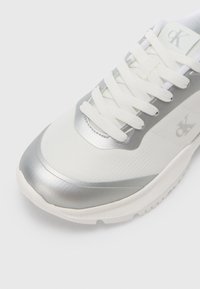 Fehér tornacipő ezüst szegélyekkel, textil felsőrész, fűzős kialakítás, lekerekített orr, és texturált talp. Logó az oldalon, minimalista dizájn.