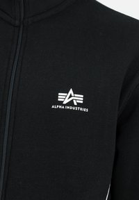 Svart zip-up hettegenser laget av mykt stoff, med en hvit logo "ALPHA INDUSTRIES" med et stilisert emblem på venstre bryst.