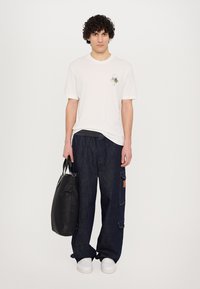 Camiseta blanca de manga corta con un pequeño gráfico, vaqueros anchos de color azul oscuro, zapatillas blancas y un bolso negro de acabado mate.