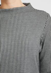 Nahaufnahme eines langärmeligen Tops mit schwarz-weißer Hahnentrittmusterung und engem, rundem Halsauschnitt, getragen von einer Person.