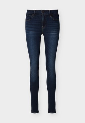 Στενά σκούρα μπλε τζιν με τσέπες μπροστά και πίσω, κλεισίματα με κουμπί και φερμουάρ, και διακριτική ξεθώριαση στους μηρούς και τα γόνατα.