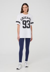Maglietta oversize bianca con la scritta "CHICAGO 93" stampata in nero. Indossata con leggings blu navy e sneaker bianche. Design semplice, maniche corte.