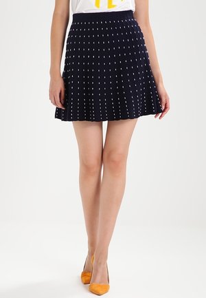 Kvinde iført en marineblå plisseret nederdel med hvide lodrette prikker og orange spidse hæle, stående mod en hvid baggrund.