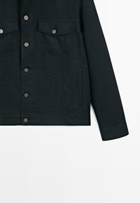 Veste en denim noire avec manches longues, boutons métalliques sur le devant, deux poches poitrine à rabats boutonnés et poches côtés passepoilées.
