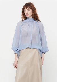 Vrouw met rood haar in een doorschijnende lichtblauwe blouse over een beige rok, die iets opzij kijkt tegen een effen witte achtergrond.