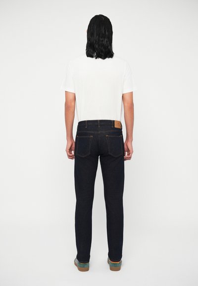 Jeans de ganga azul escuro com um design ajustado, apresentando costuras contrastantes, bolsos traseiros e uma etiqueta de couro castanho na cintura.
