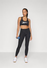 Črna športna modrc s belim logotipom, visoko pasovi črne legice in bele športne čevlje. Drži modro utež. Gladka tekstura.