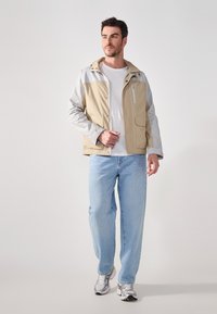 Beige og grå lett jakke med frontglidelås, sidelommer og justerbar hette, sammen med lyseblå jeans og sølvfargede joggesko.