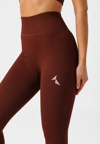 Vysoké sportovní legíny v bohaté hnědé ribbované textuře, se malým bílým logem na spodní části nohy.