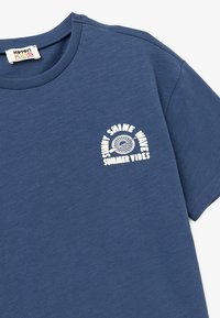Camiseta de algodón azul con cuello redondo y mangas cortas. El diseño en la parte frontal izquierda incluye texto blanco y un gráfico circular.