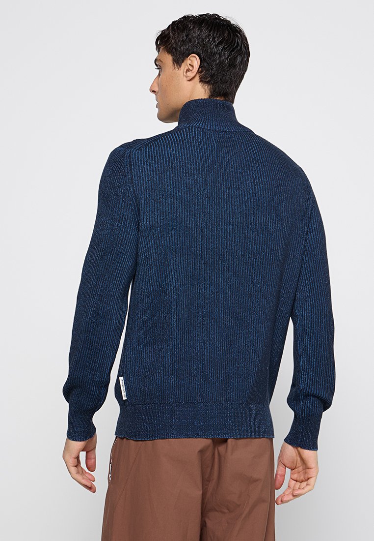 Mann in einem gerippten navyblauen Rollkragenpullover und braunen Hosen, der sich von einer einfachen hellen Hintergrund abwendet.