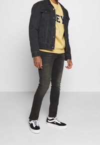 Black denim jacket over a yellow sweatshirt with blue lettering, paired with dark jeans and black and white sneakers.