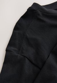 Gros plan sur un tissu noir montrant une couture détaillée le long d'une couture de manche sur un vêtement posé à plat sur une surface claire.