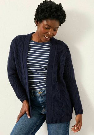 Navy knitted cardigan with a textured pattern and open front, worn over a navy and white striped shirt and blue jeans.
