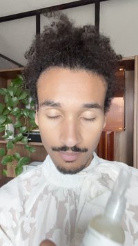 Personne calme avec des cheveux bouclés et une moustache, portant un haut en dentelle blanche, tenant un flacon pulvérisateur dans une pièce avec des étagères en bois et une plante verte.