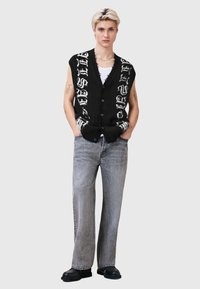 Melns piedurkņu kardigans ar baltu grafisku tekstu, priekšpusē ar pogām, kopā ar pelēnām platajiem džinsiem un melniem apaviem. Minimālistisks stils.