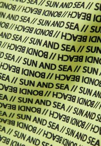 Close-up van stof met herhaalde zwarte tekst "SUN AND SEA // BONDI BEACH" op een lichtgroene achtergrond.