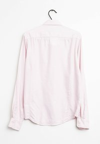 Camicia rosa chiaro a maniche lunghe con colletto a punta. Presenta un orlo dritto, polsini con un bottone e una texture liscia. Appesa a un gancio nero.