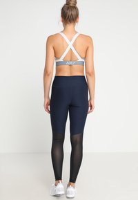 Szary stanik sportowy z krzyżującymi się ramiączkami i logo Nike, w połączeniu z granatowymi legginsami z siateczkowymi wstawkami oraz białymi butami sportowymi.