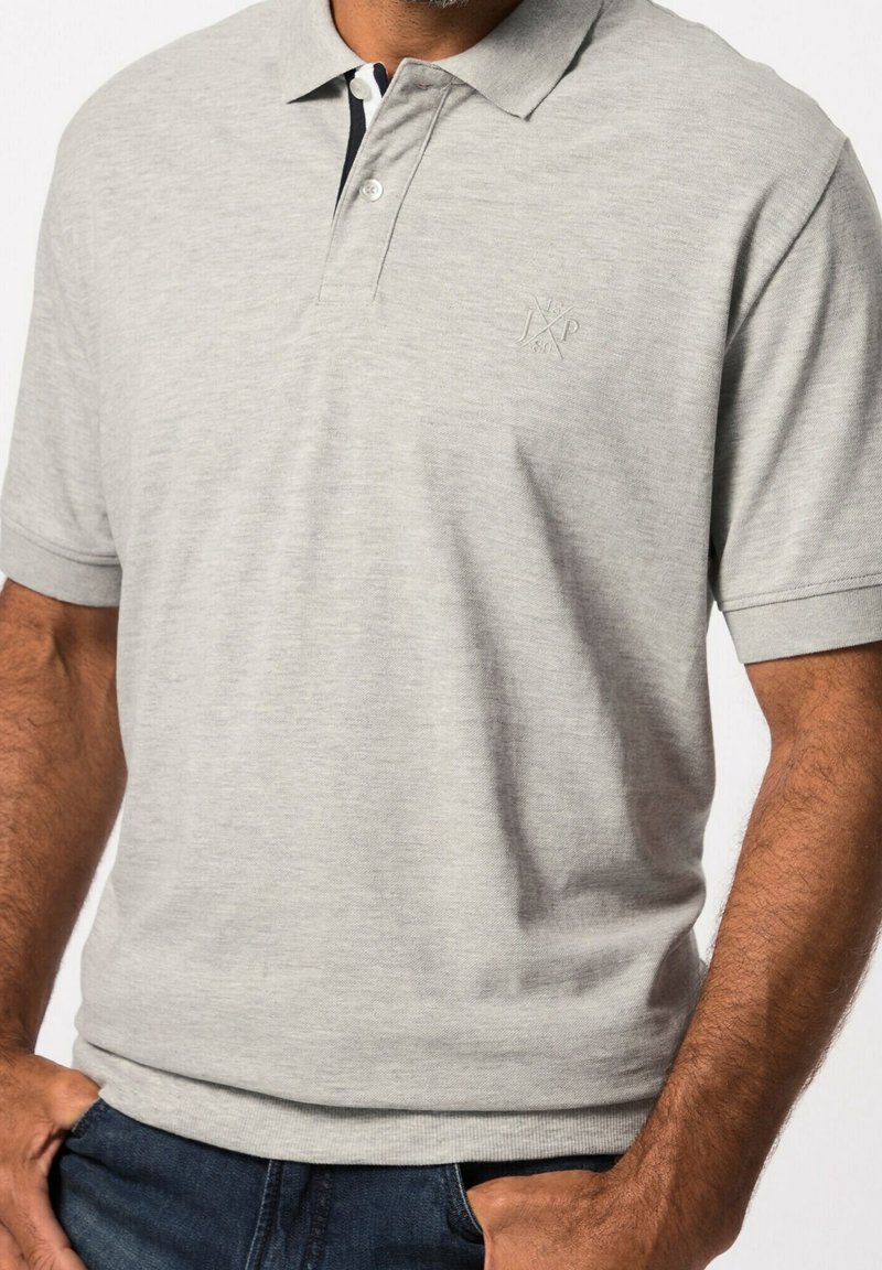 Homme portant un polo gris clair à manches courtes et un jean bleu foncé, mains partiellement dans les poches, aucun arrière-plan visible.