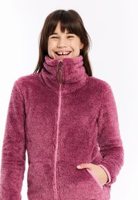 Nasmejana djevojka s dugom smeškom kose u roza dukserici s patentnim zatvaračem, s desnom rukom u džepu dukserice, naspram bijele pozadine.