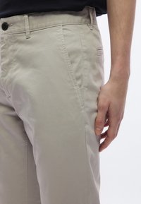 Béžové bavlnené nohavice so strečovým strihom, vybavené bočnými vreckami, pútkami na opasok a jedným zadným vreckom so zapínaním na gombík.