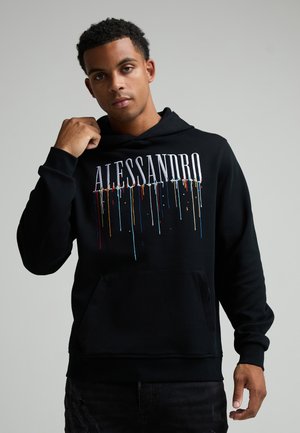 Jeune homme aux cheveux courts et bouclés portant un sweat à capuche noir avec le texte "ALESSANDRO" et un design coloré de gouttes de peinture, ajustant la capuche.