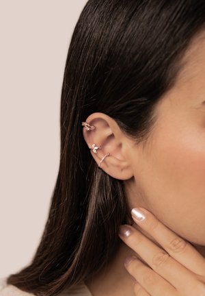 Frauenohr mit drei Roségold-Ohrringen: ein gedrehter Reifen am oberen Ohr, ein kleiner Kristall-Cluster in der Mitte und ein dünnes Band am Ohrläppchen.